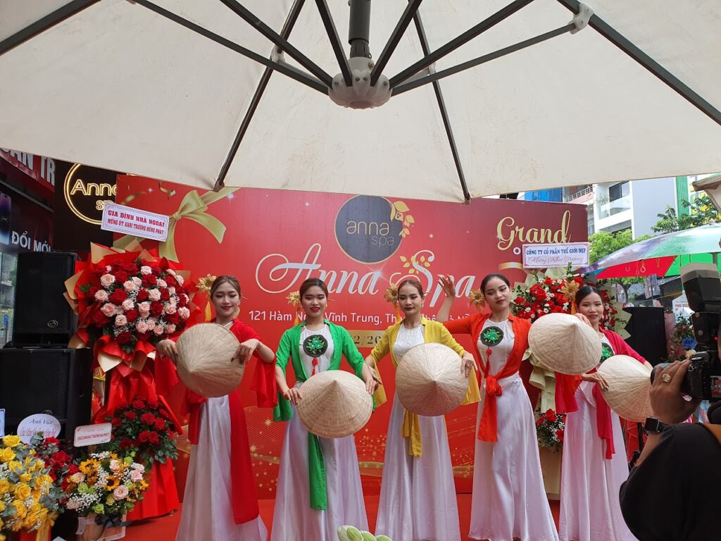 베트남 전통 의상인 아오자이와 농라(원뿔형 모자)를 들고 서 있는 다섯 명의 여성들. 여성들은 흰색 아오자이 위에 초록색, 노란색, 빨간색의 긴 천을 두르고 포즈를 취하고 있다. 오른쪽 아래에는 이 장면을 촬영하는 사람의 모습이 일부 보인다.