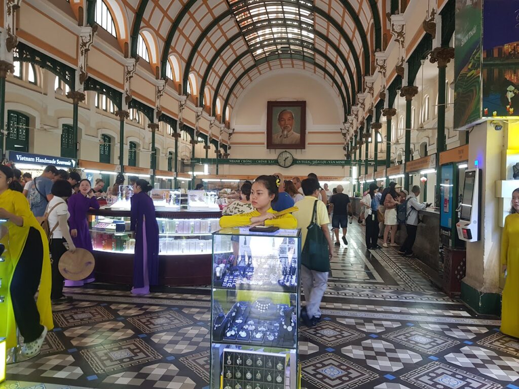 웅장한 아치형 천장을 가진 호치민 중앙 우체국 내부에서 베트남 전통 의상인 아오자이를 입은 여성들이 기념품 판매대를 구경하고 있습니다. 이 건물은 호치민 비행기표를 끊고 도착한 여행객들이 방문하는 주요 명소입니다.