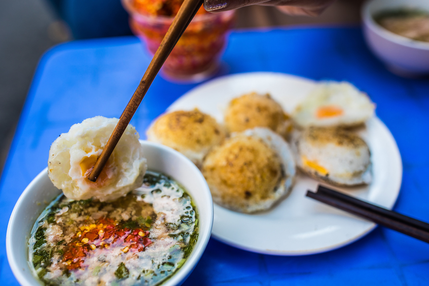 젓가락으로 잡은 흰색 쌀 케이크(반 베오 또는 유사한 음식) 조각을 고추, 쪽파, 그리고 향신료가 떠 있는 묽은 갈색 소스(액젓 기반)가 담긴 작은 그릇에 찍고 있는 모습. 배경의 파란색 탁자 위 흰 접시에는 비슷한 모양의 노르스름한 케이크 몇 조각이 놓여 있습니다. (베트남 길거리 음식인 반 베오나 반콧으로 보임)