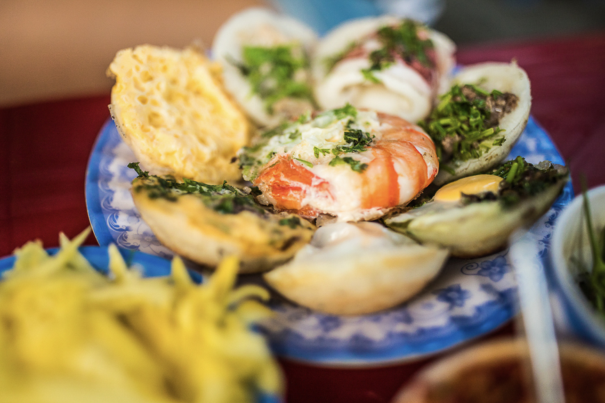 접시에 담긴 베트남 거리 음식 ‘반껀(Bánh Căn)’으로, 조개껍데기 모양 틀에 구운 작은 전 위에 새우, 달걀, 파 등이 올려져 있는 모습이 클로즈업으로 보인다.