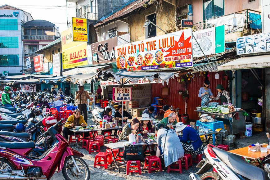 달랏의 번화가 노점에서 사람들이 플라스틱 테이블과 빨간 의자에 앉아 현지 음식을 먹고 있는 모습. 주변에는 오토바이들이 주차되어 있고, 베트남어 간판이 즐비하다.