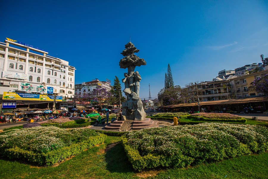 베트남 달랏 중앙 시장 앞 광장 전경. 중앙에는 독특한 모양의 조각상 기념비가 서 있고, 주변에는 잘 가꿔진 꽃밭과 푸른 잔디가 펼쳐져 있다. 배경으로는 왼쪽에는 현대적인 호텔 건물과 시장 노점들이, 오른쪽에는 달랏 시내의 건물들이 보인다. 맑고 푸른 하늘이 인상적이다.