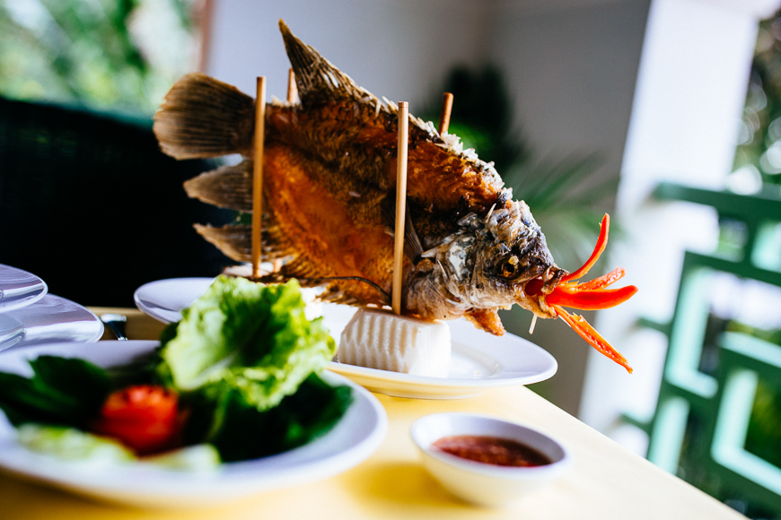 껀터 여행에서 맛볼 수 있는 메콩 델타의 특별한 요리인 바삭하게 튀겨낸 생선 (Slap-Slap fish / Elephant Ear Fish) 요리입니다. 생선이 접시에 세워져 있고, 채소와 소스가 함께 제공됩니다.