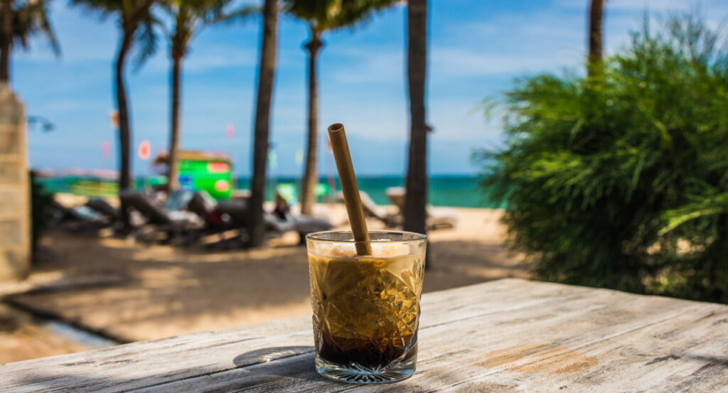 야자수가 드리워진 해변을 배경으로 나무 테이블 위에 놓인 시원한 아이스 커피 한 잔의 클로즈업 사진입니다. 무이네 숙소 싸게 잡고 휴식을 취하며 즐기는 베트남 커피의 여유.