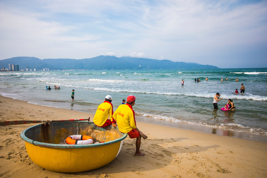 잔잔한 파도가 밀려오는 바다에는 여러 명의 관광객들이 물놀이를 즐기고 있으며, 일부는 튜브나 작은 보드를 사용하고 있습니다. 배경에는 높은 건물들이 늘어선 다낭 시내와 푸른 산맥이 길게 펼쳐져 있어, 해변이 다낭 당일치기 또는 여행의 주요 코스로 적합함을 보여줍니다.