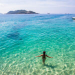 맑고 푸른 바다에서 한 여성이 서서 물놀이를 즐기고 있는 전경. 수면 아래 모래까지 투명하게 보이는 나트랑여행의 아름다운 해변.