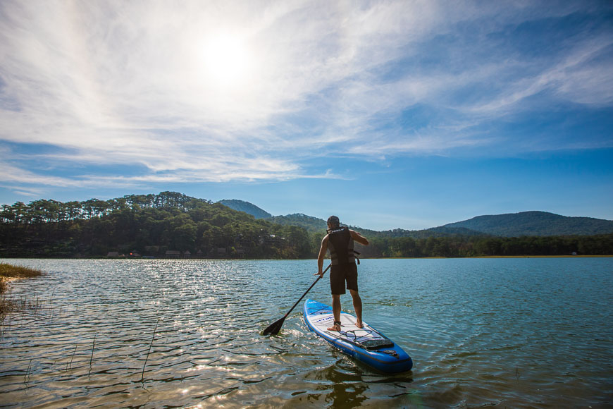맑고 푸른 하늘 아래, 한 남성이 잔잔한 호수 위에서 패들보드를 타고 있는 모습. 배경에는 소나무가 우거진 산들이 펼쳐져 있다.
