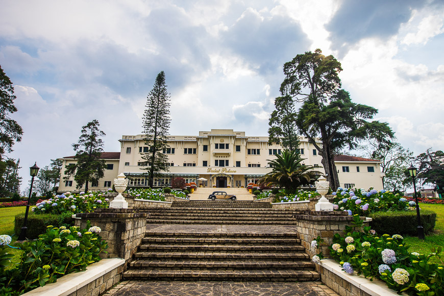 Dalat Palace'라는 이름이 새겨진 중앙 입구와 여러 층으로 이루어진 크림색의 웅장한 콜로니얼 양식 호텔 건물 전경. 건물의 긴 계단 아래에는 돌담과 화단이 있으며, 화단에는 파스텔 톤의 **수국(Hydrangea)**이 가득 피어 있습니다. 계단 중앙에는 클래식한 모양의 어두운색 자동차 한 대가 주차되어 있고, 양옆으로는 키 큰 침엽수와 활엽수가 우뚝 솟아 있습니다. 하늘은 구름이 많습니다. (베트남 달랏 팰리스 호텔)