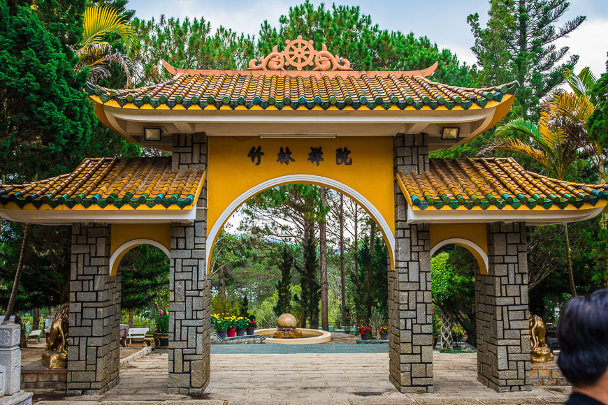 노란색 벽과 갈색 기와 지붕이 특징인 베트남 전통 건축 양식의 사찰 입구(산문). 문 중앙 상단에는 竹林禪院(죽림선원)이라는 한자가 새겨져 있으며, 지붕 위에는 불교 상징인 황금색 수레바퀴 모양 장식이 있습니다. 문은 벽돌 무늬의 돌 기둥으로 이루어져 있고, 아치형 통로 너머로는 울창한 소나무와 정원이 보입니다. (베트남 달랏의 쯕럼 선원(Truc Lam Zen Monastery) 입구)