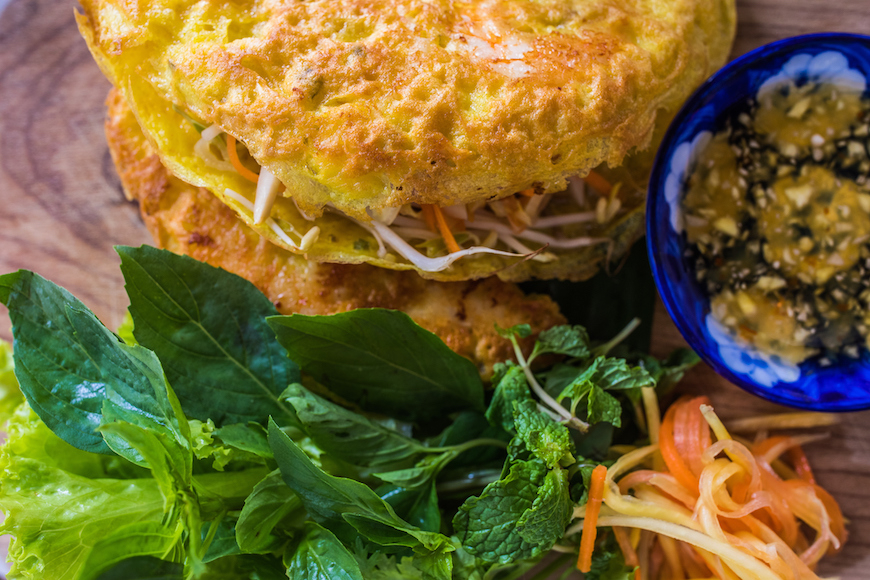 베트남 중부의 길거리 음식인 반쎄오(Bánh Xèo)의 클로즈업 사진. 노란색의 바삭한 부침개 속에는 숙주와 새우 등이 들어 있으며, 옆에는 상추, 민트, 베트남 허브 등의 신선한 채소와 당근 채가 담겨 있다. 오른쪽에는 작은 파란색 그릇에 달콤하고 짭짤한 땅콩 소스(느억 짠)가 담겨 있다.
