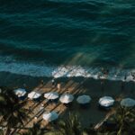 초록빛 바다가 보이는 해변에서 여러 명의 사람들이 파도 근처에서 물놀이를 하고 있다. 해변에는 하얀 파라솔과 선베드가 줄지어 놓여 있으며, 야자수의 그림자가 길게 드리워져 있다