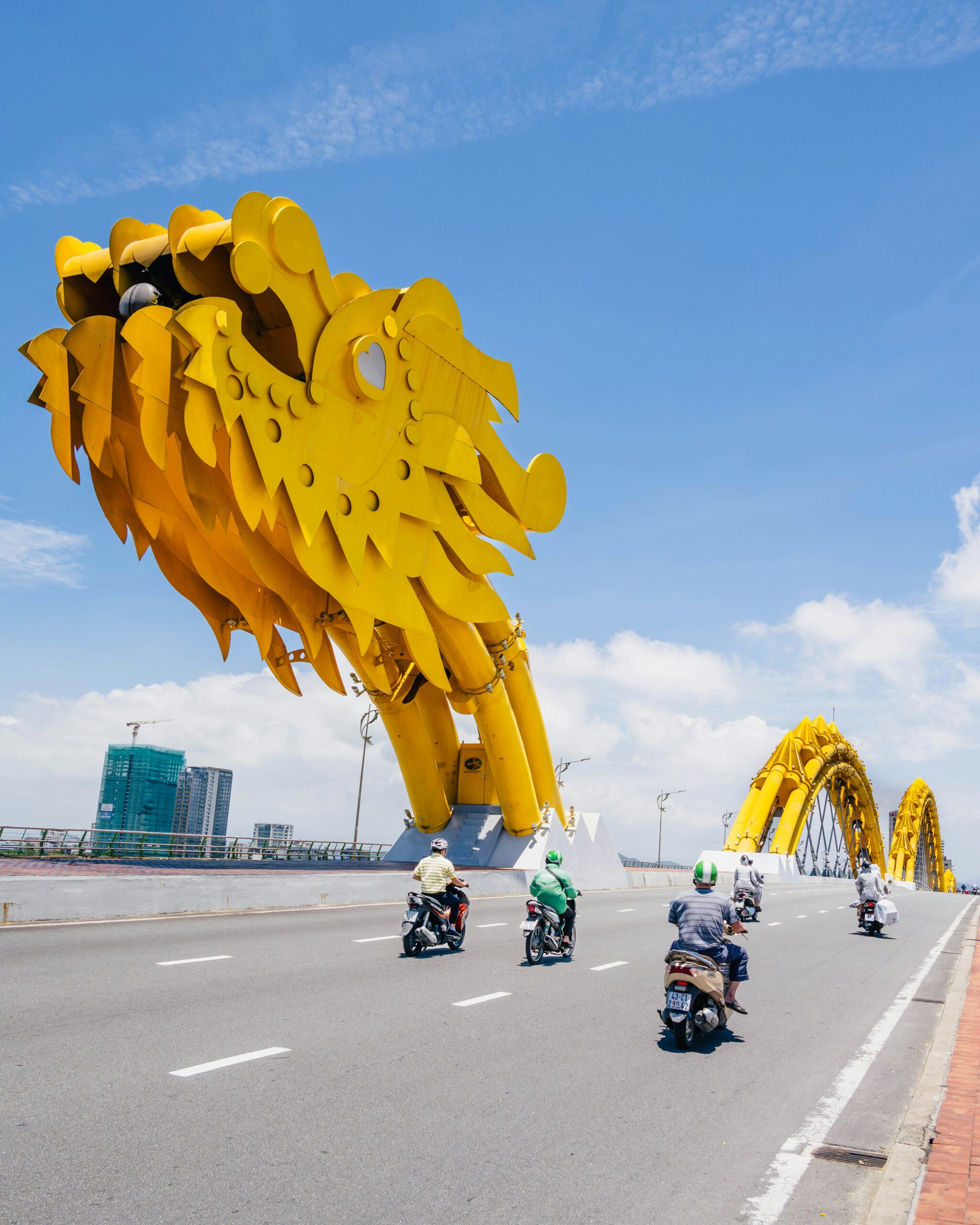 푸른 하늘과 흰 구름 아래, 다낭 용다리(Dragon Bridge) 위를 달리는 오토바이들의 모습. 다리 위에 설치된 거대한 황금색 용 머리 구조물이 시선을 압도하며, 뒤쪽으로는 다리의 아치 구조물과 시내 고층 건물들이 보인다. 차도는 밝고 깨끗한 상태이다.