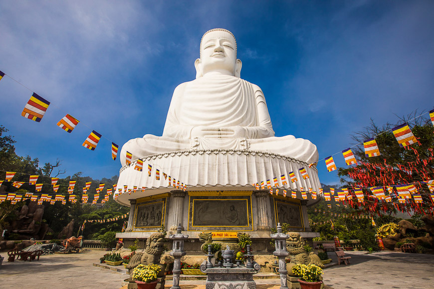 다낭의 영흥사(Linh Ung Pagoda)에 위치한 거대한 흰색 불상(대좌불)의 모습. 불상은 연화대 위에 좌선한 자세로 앉아 있으며, 주변에는 베트남 불교의 상징인 오색 깃발들이 일렬로 걸려 있다. 파란 하늘을 배경으로 넓은 광장과 불상을 장식한 기단부가 보인다.