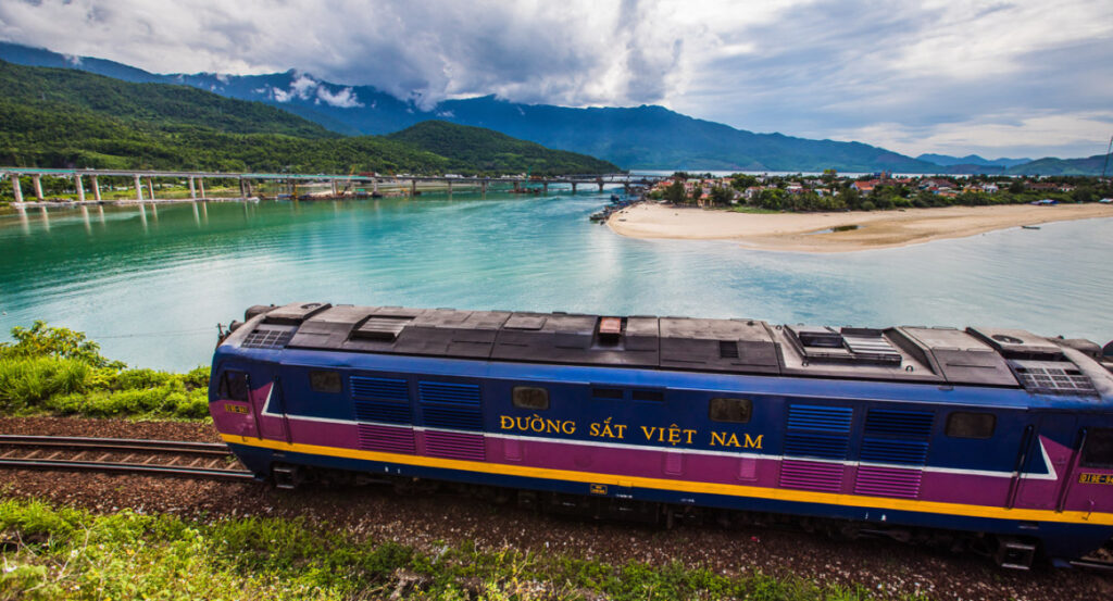 베트남의 아름다운 해안선을 따라 달리는 파란색 기차의 모습입니다. 에메랄드빛 바다와 산맥이 어우러진 풍경이 인상적이며, 무이네 싸게 가기를 위한 기차 여행의 매력을 보여줍니다.