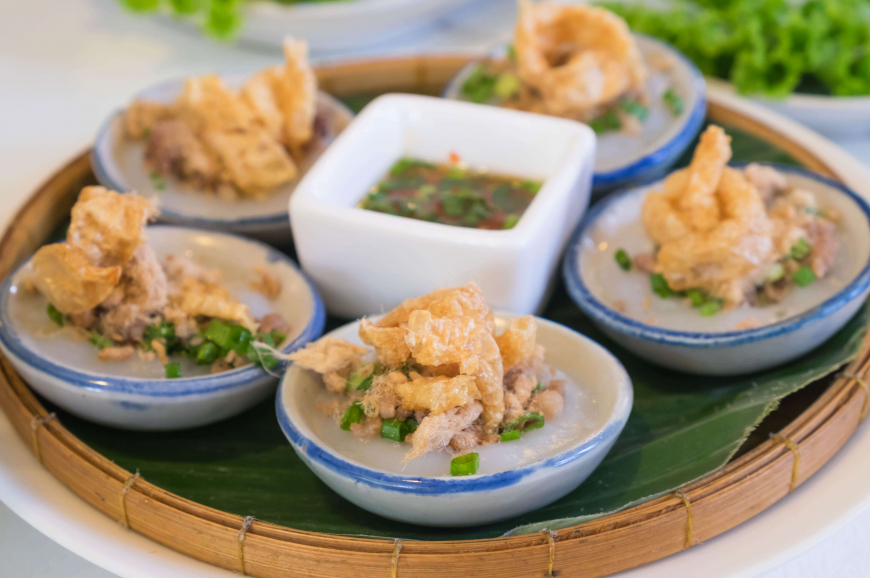 바나나 잎 위에 담긴 베트남 음식 반베오(Bánh Bèo). 작은 접시에 담긴 쌀가루 케이크 위에 새우와 돼지고기 토핑, 그리고 튀긴 돼지 껍질이 얹혀 있습니다. 이 음식은 호치민 비행기표를 끊고 온 여행자들이 현지에서 즐기는 다양한 미식 중 하나입니다.
