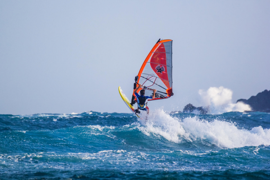무이네 당일치기 중 해변에서 즐기는 윈드서핑 액티비티. 서퍼가 파도를 타고 힘차게 뛰어오르는 역동적인 순간을 담고 있으며, 오렌지색 돛이 눈에 뜁니다.