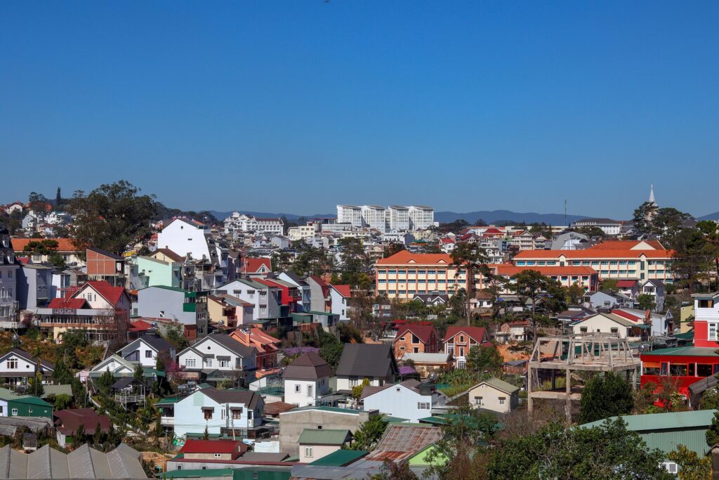 맑고 푸른 하늘 아래 언덕에 자리 잡은 베트남 달랏의 주택가 전경. 다양한 색상과 건축 양식의 집들이 밀집해 있으며, 일부 현대적인 큰 건물도 보입니다. (베트남 달랏 시가지 전경)