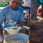 껀터 당일치기 여행 중, 작은 배 위에서 현지인이 갓 삶은 쌀국수를 그릇에 담아 내는 모습. 따뜻한 국물에서 김이 오르고, 주변에는 재래 수상시장의 풍경이 보인다.