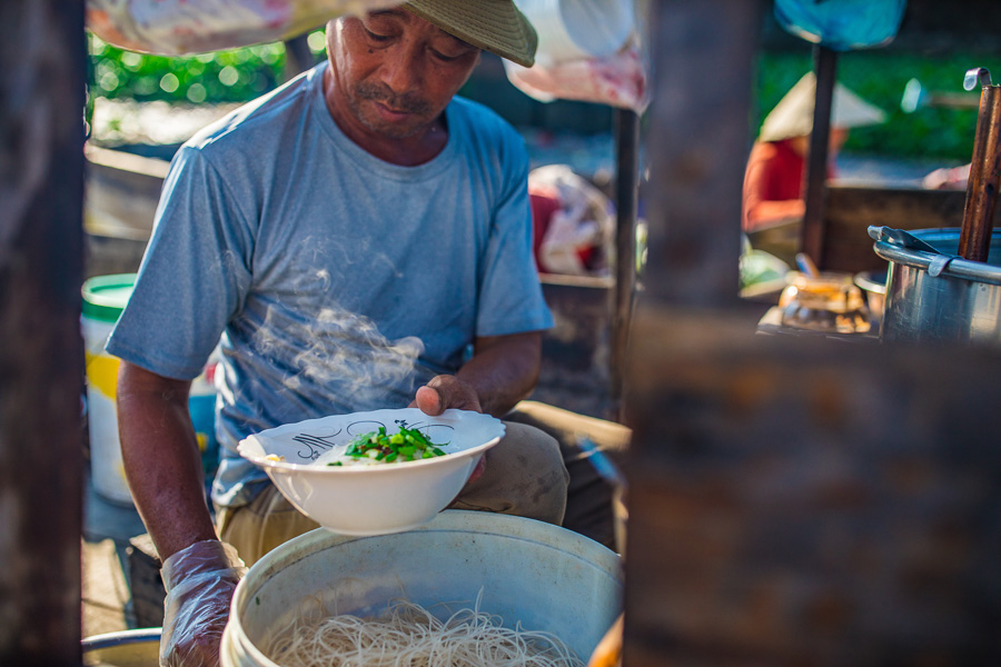 껀터 당일치기 여행 중, 작은 배 위에서 현지인이 갓 삶은 쌀국수를 그릇에 담아 내는 모습. 따뜻한 국물에서 김이 오르고, 주변에는 재래 수상시장의 풍경이 보인다.