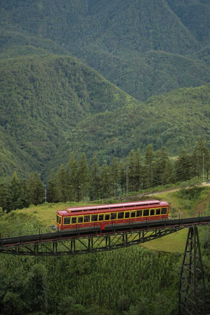 울창한 녹색 산맥과 숲을 배경으로, 붉은색과 노란색의 관광 열차(푸니쿨라 또는 트램)가 검은색 철교 위를 가로지르는 모습. 이 이미지는 베트남 사파의 아름다운 자연경관과 여행 분위기를 보여주며, 인근 사파 숙소에서 볼 수 있는 풍경을 연상시킵니다.