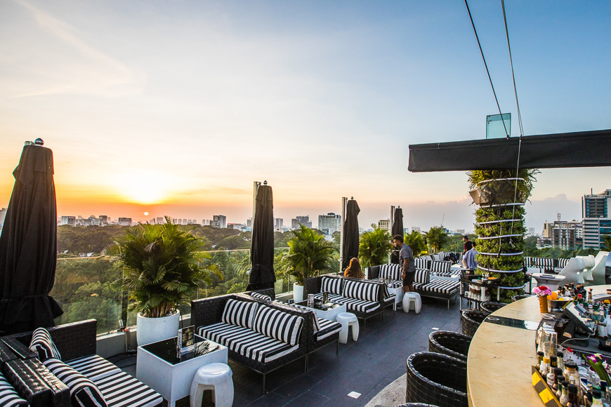호치민 도심의 루프탑 바에서 찍은 일몰 풍경. 도시 스카이라인을 배경으로 해가 지고 있으며, 바의 테라스에는 줄무늬 소파와 식물이 배치되어 휴식을 즐기는 사람들이 보입니다.
